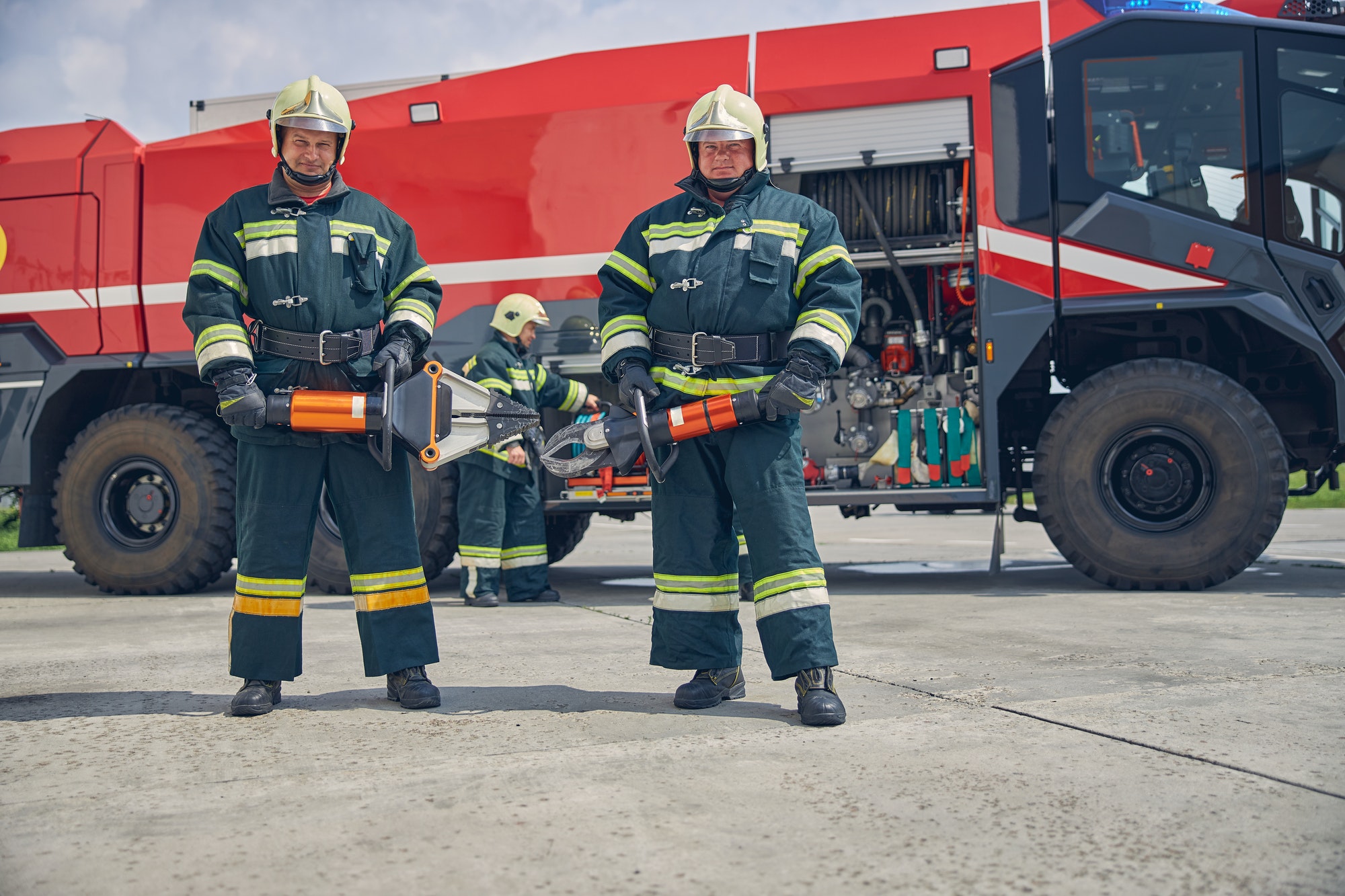 Strong handsome firefighters standing near the fire machine with equipment in hands